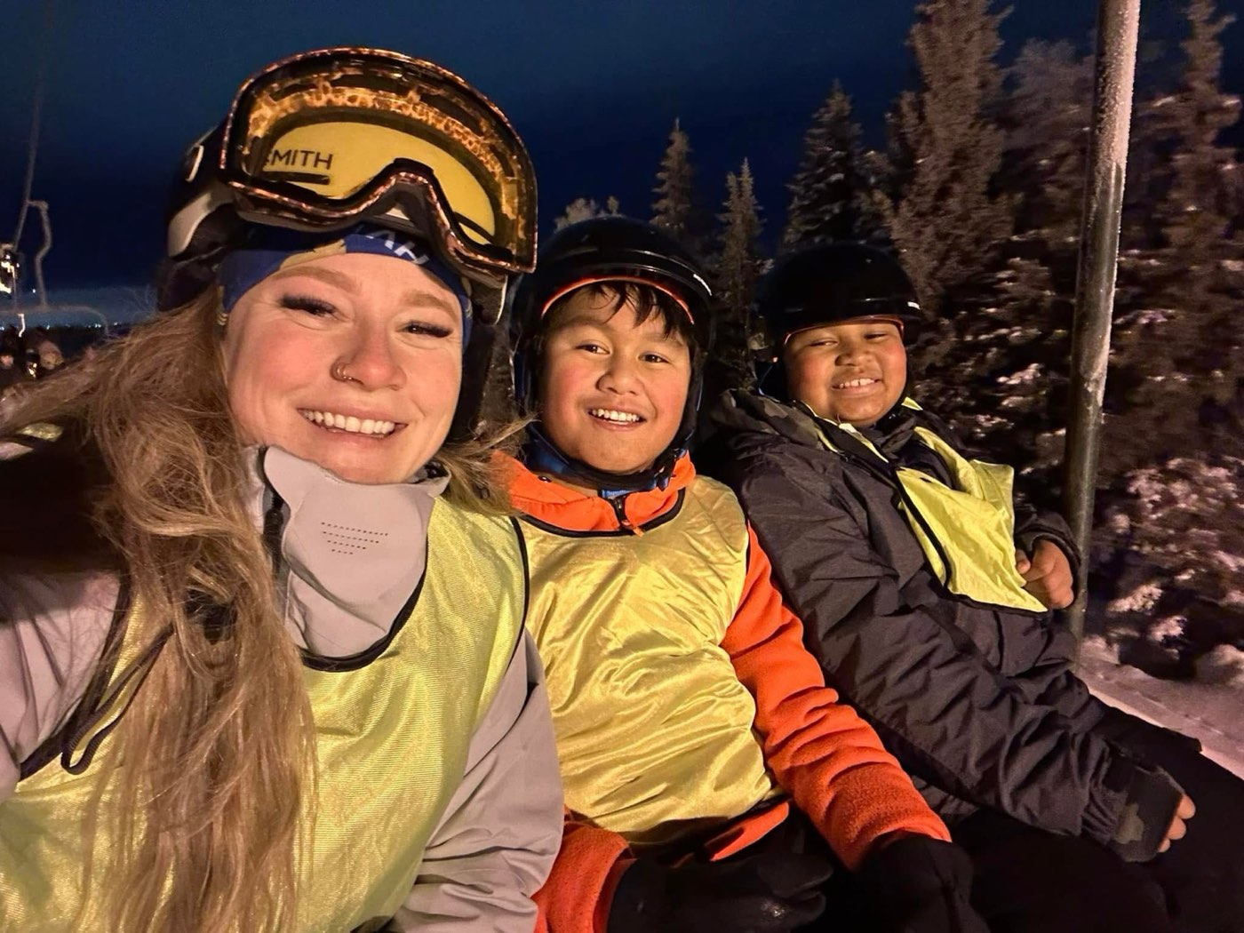 A mentor and two kids smiling on the chairlift during a night ski session at Hilltop.