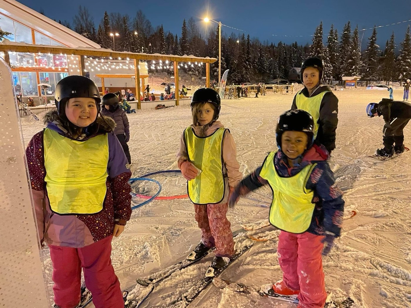 Four kids in yellow lesson bibs smiling at the lodge before a night ski session.