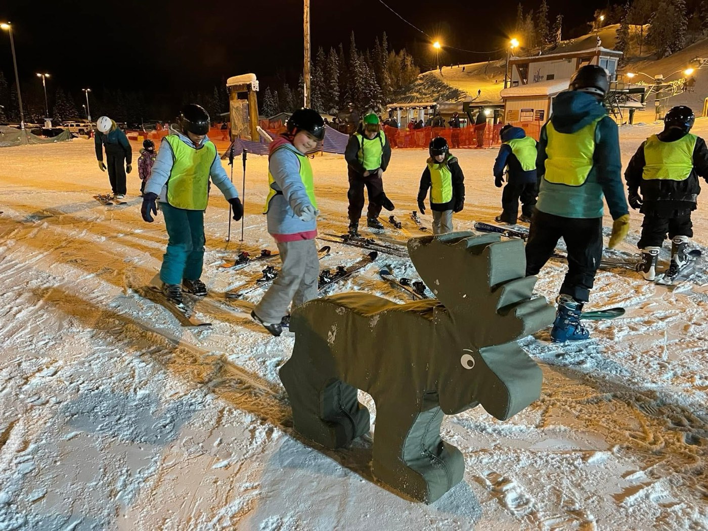 Kids in yellow lesson bibs gathered around the Hilltop moose mascot on the snow at night.